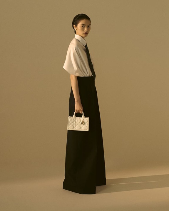 Person wearing a formal outfit holding a small white purse, standing against a beige background.