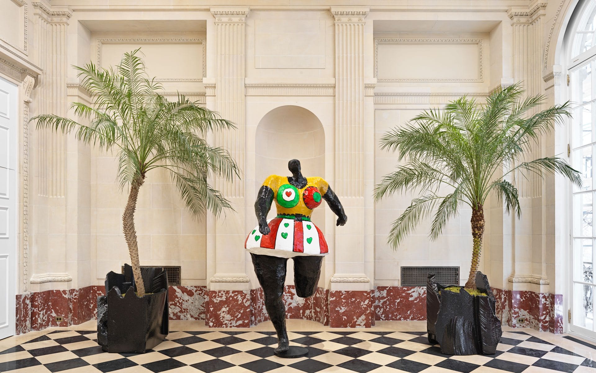 Colorful sculpture of a woman in a dress, flanked by two potted palm trees, set against a classical architectural backdrop.