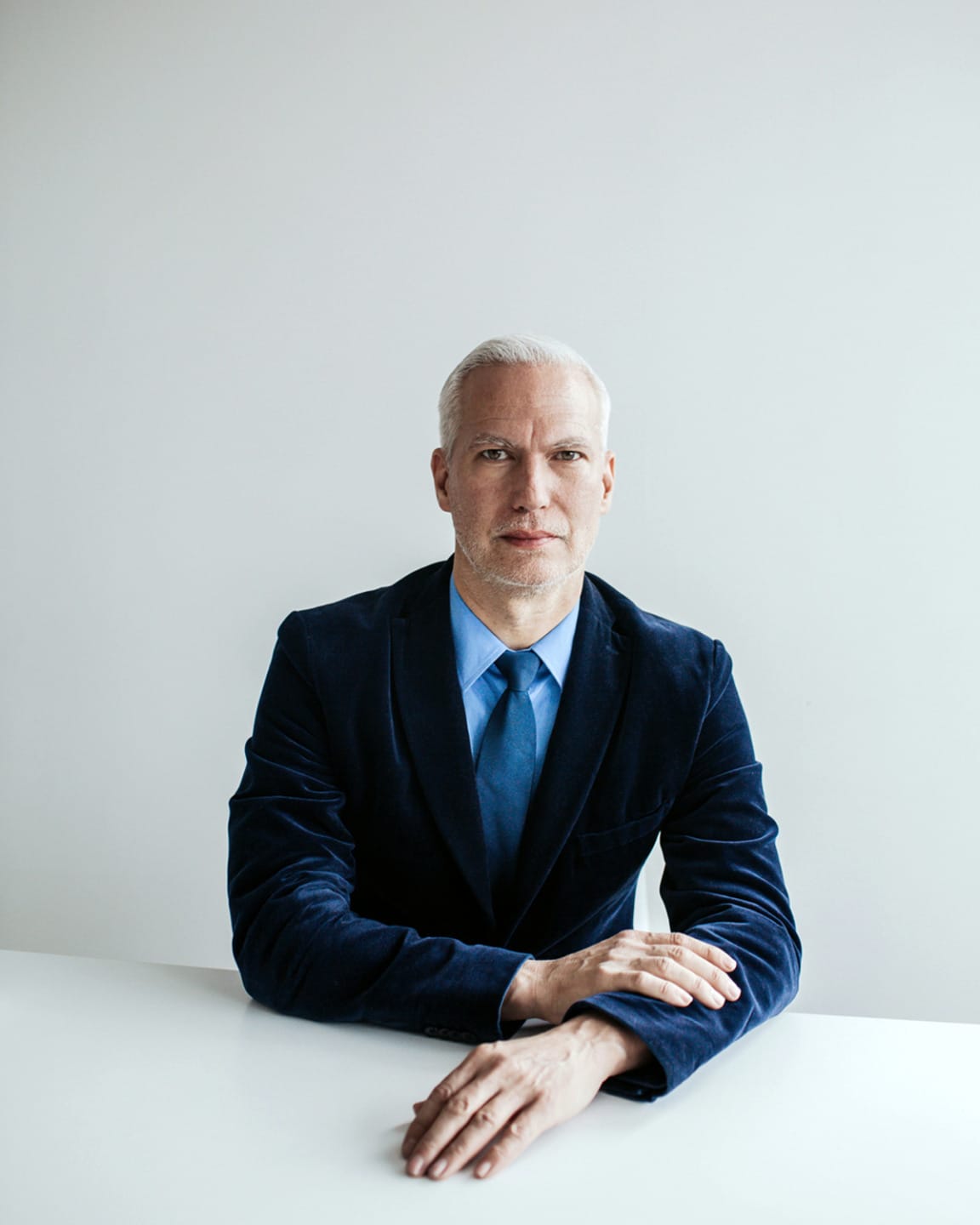 Man in a blue suit seated at a white table against a plain background.
