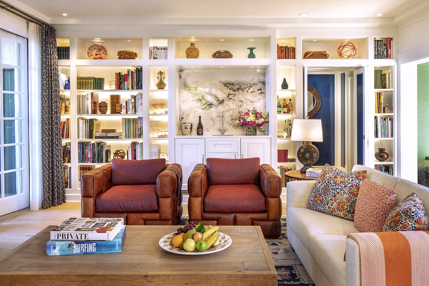 Cozy living room with orange armchairs, decorative pillows on beige sofa, and bookshelves filled with books and decor items.