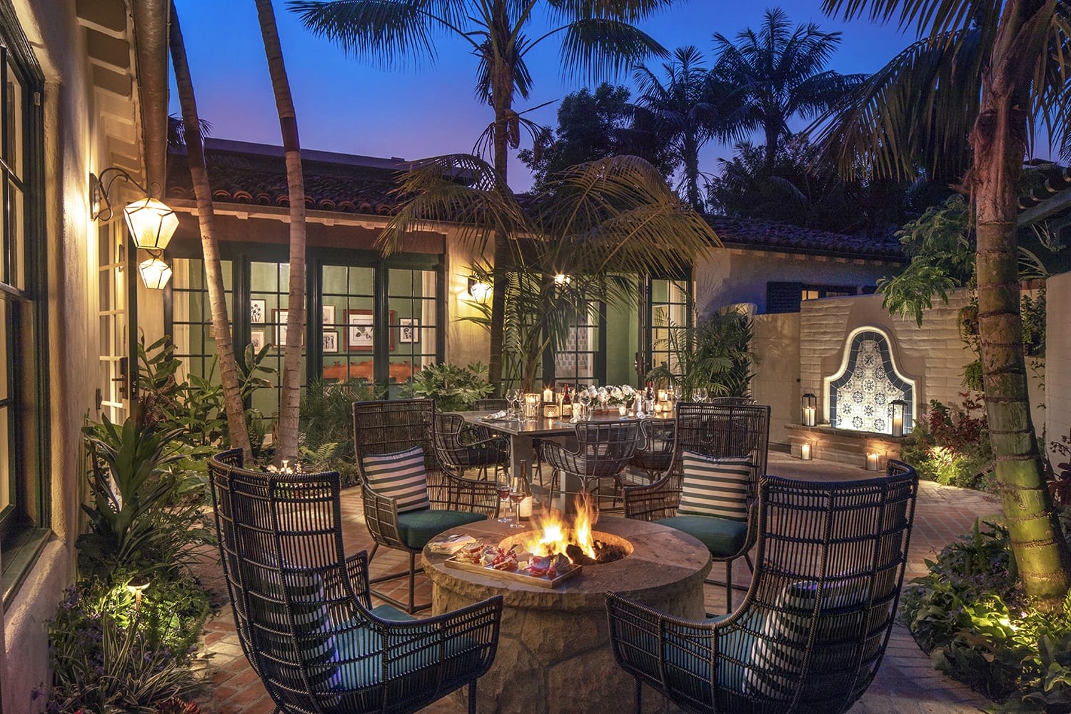 Cozy patio with a fire pit, surrounded by wicker chairs, decorated with palm trees and ambient lighting at dusk.