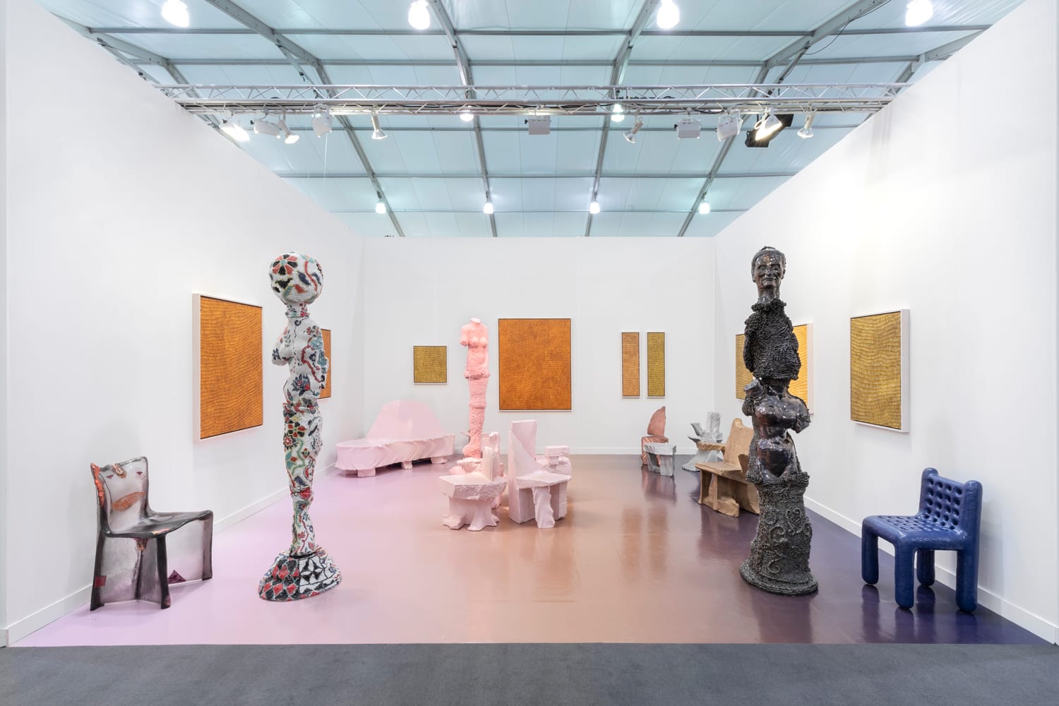 Contemporary art exhibit featuring abstract sculptures, colorful chairs, and textured paintings on white walls in a gallery setting.