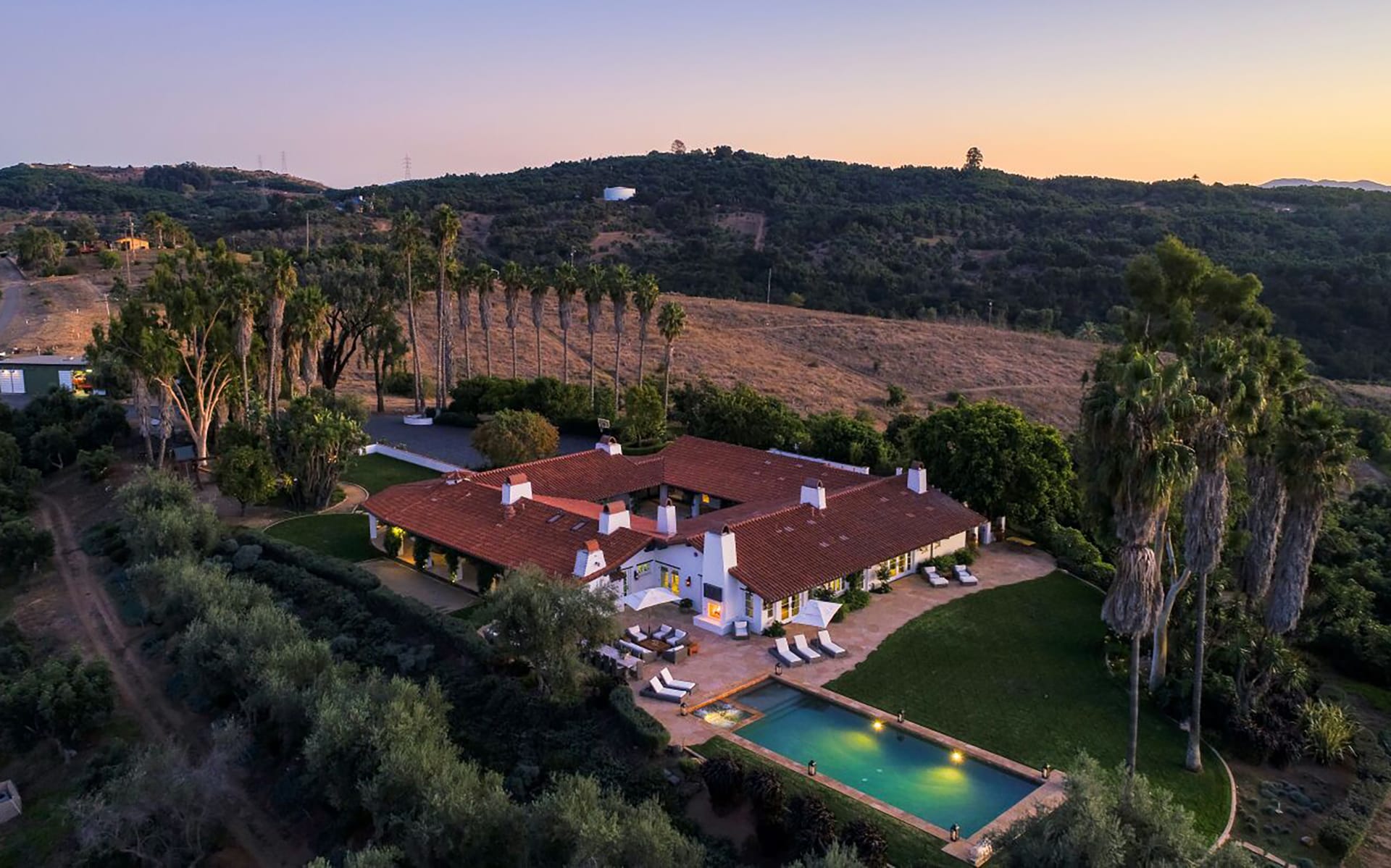 Aerial view of a Mediterranean-style villa with a red-tiled roof, pool, and surrounding trees at sunset.