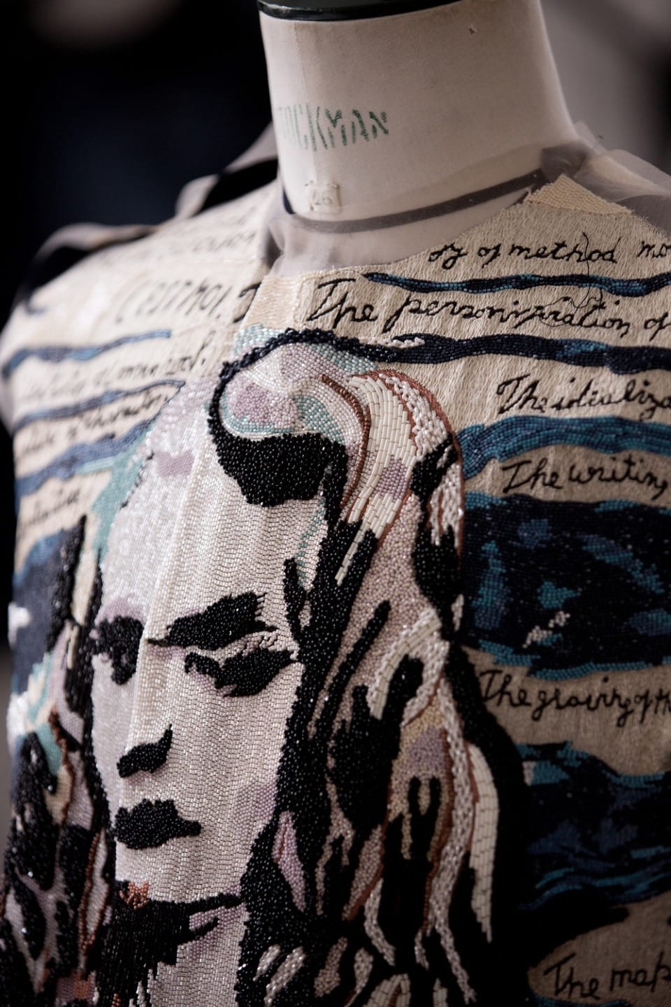 Dress with a beaded portrait and handwritten text displayed on a mannequin showing intricate artistic detail.