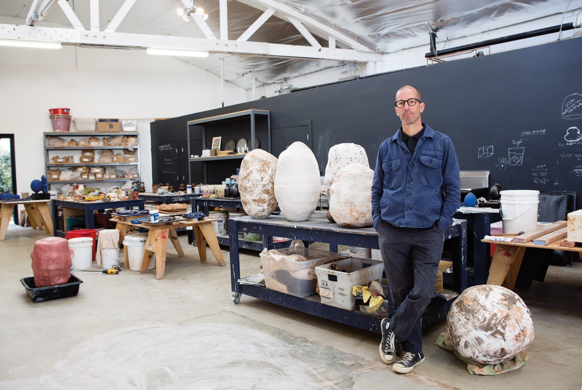 Person standing in an art studio with unfinished pottery and tools on workbenches in the background.