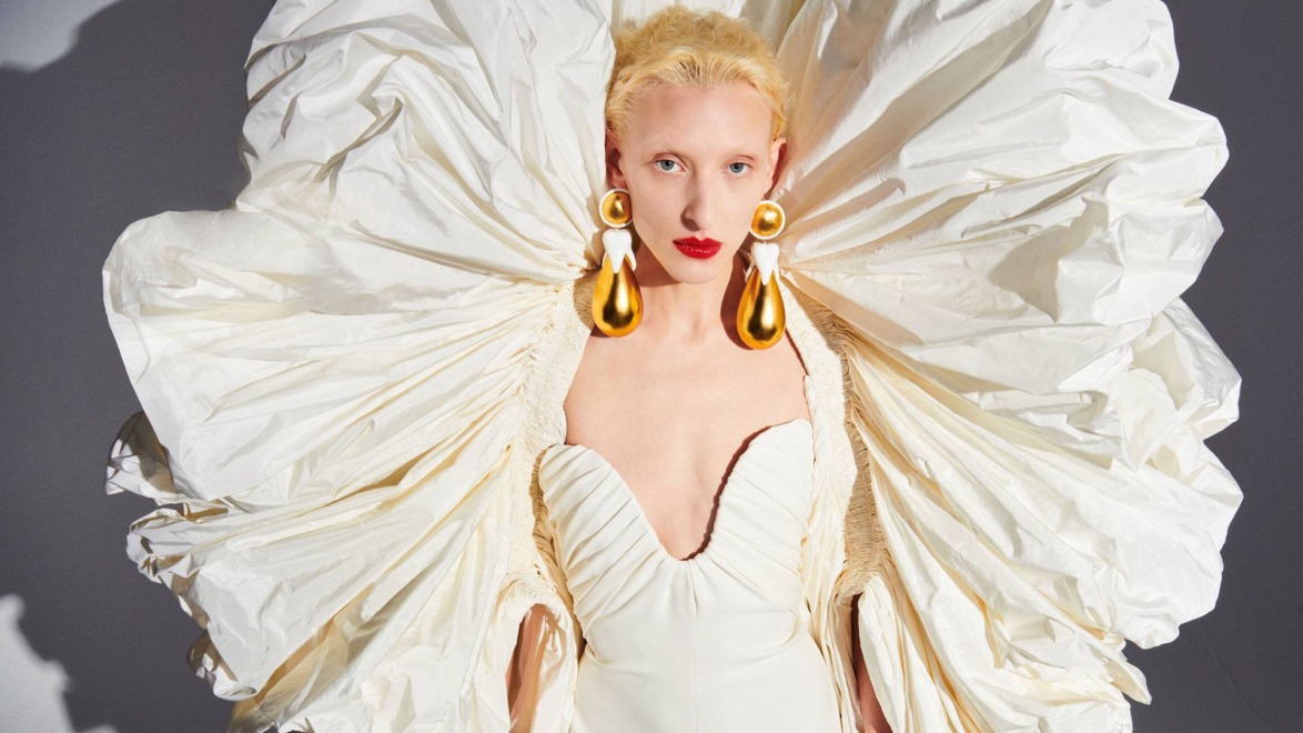 Fashion model in an elaborate white gown with large gold earrings and bold red lipstick against a grey background.