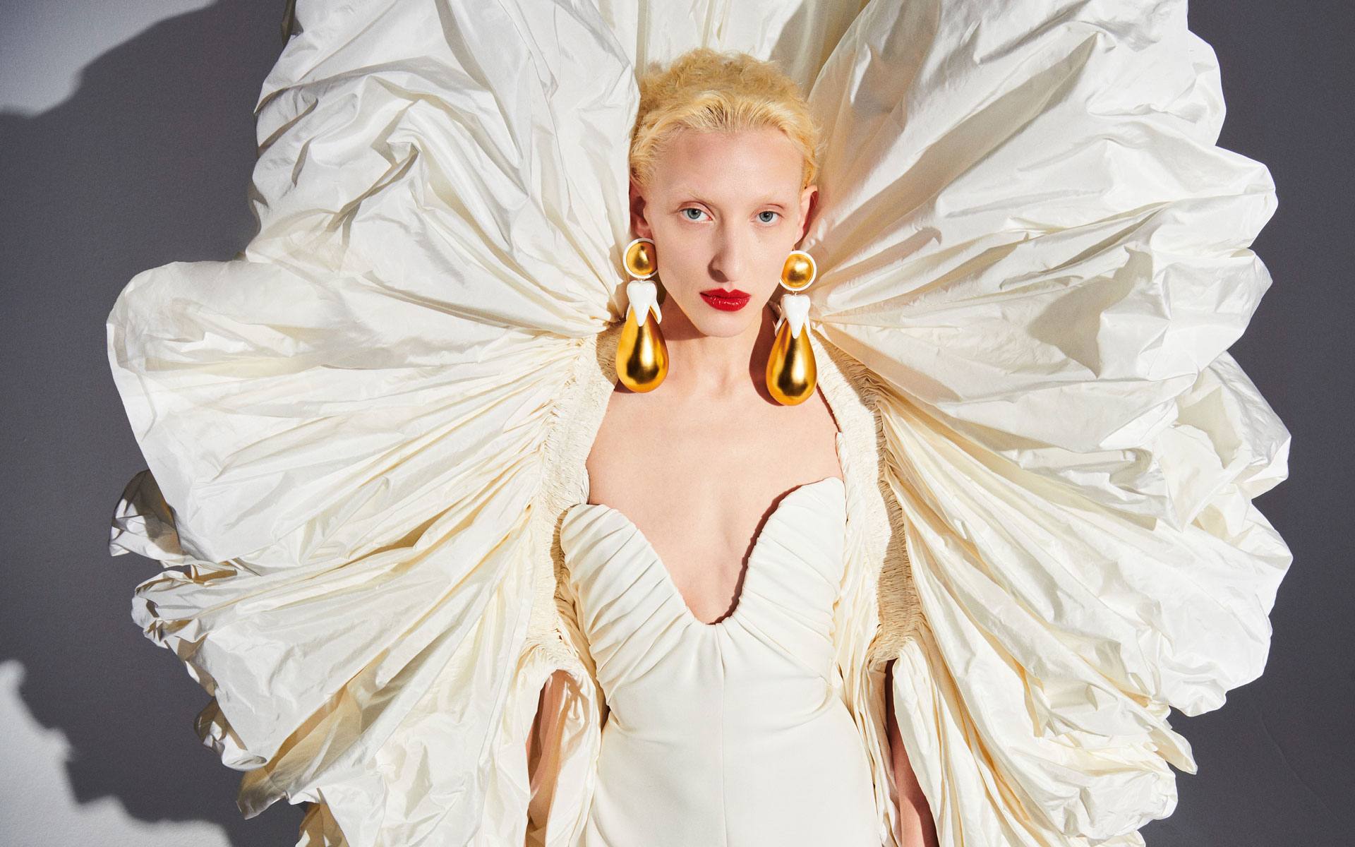 Fashion model in an elaborate white gown with large gold earrings and bold red lipstick against a grey background.