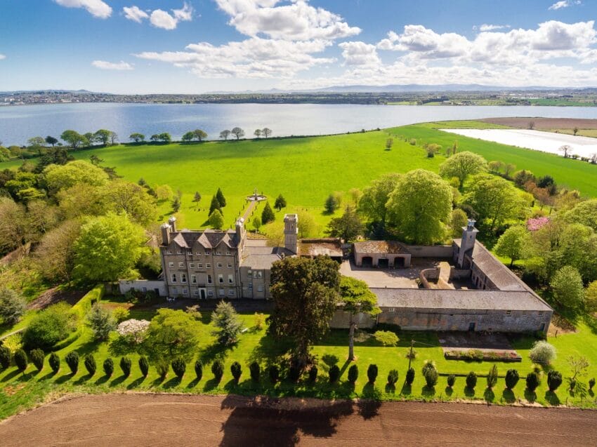 Aerial view of a historic stone castle surrounded by lush green fields and a scenic lake in the background.