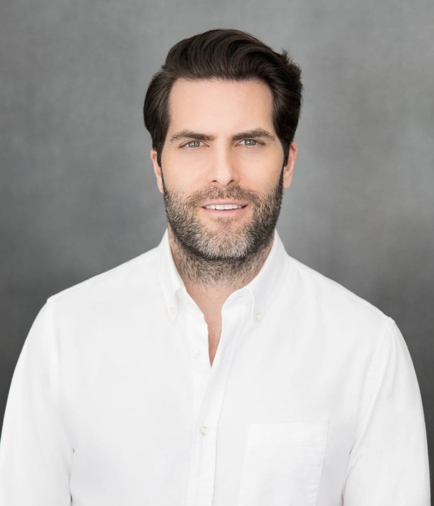 Man in a white shirt with short hair and a beard, standing against a gray background.