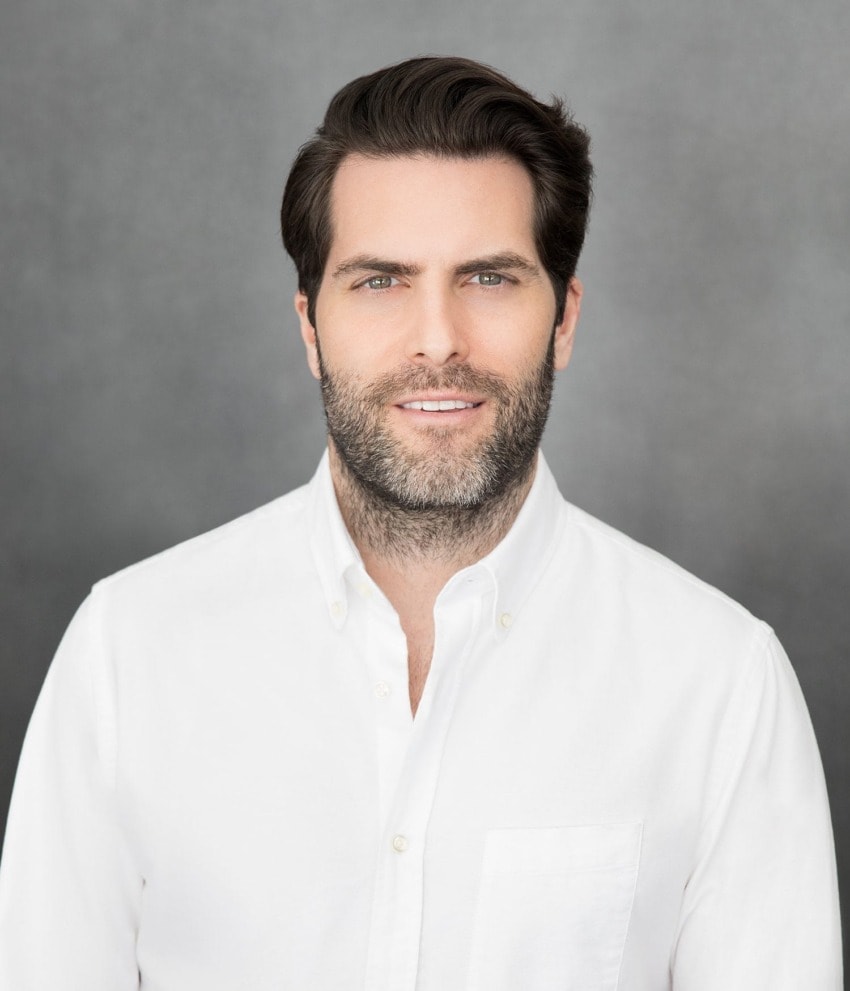 Man in a white shirt with short hair and a beard, standing against a gray background.