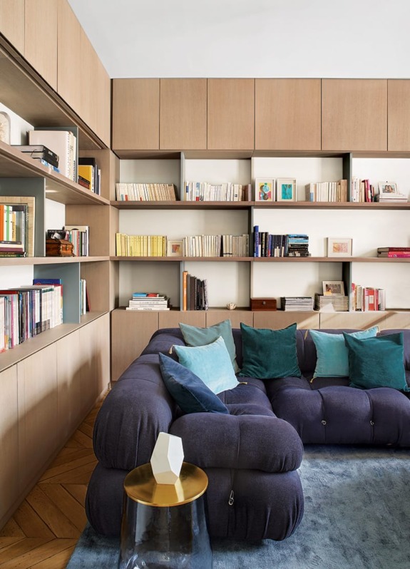 Modern living room with a blue sectional sofa, colorful pillows, wooden bookshelves, and a small round table.