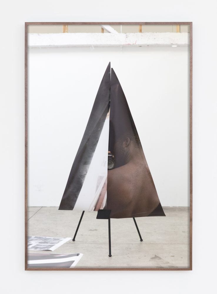 Abstract artwork with triangular shape and partial view of a person, displayed in a frame against a white gallery wall.