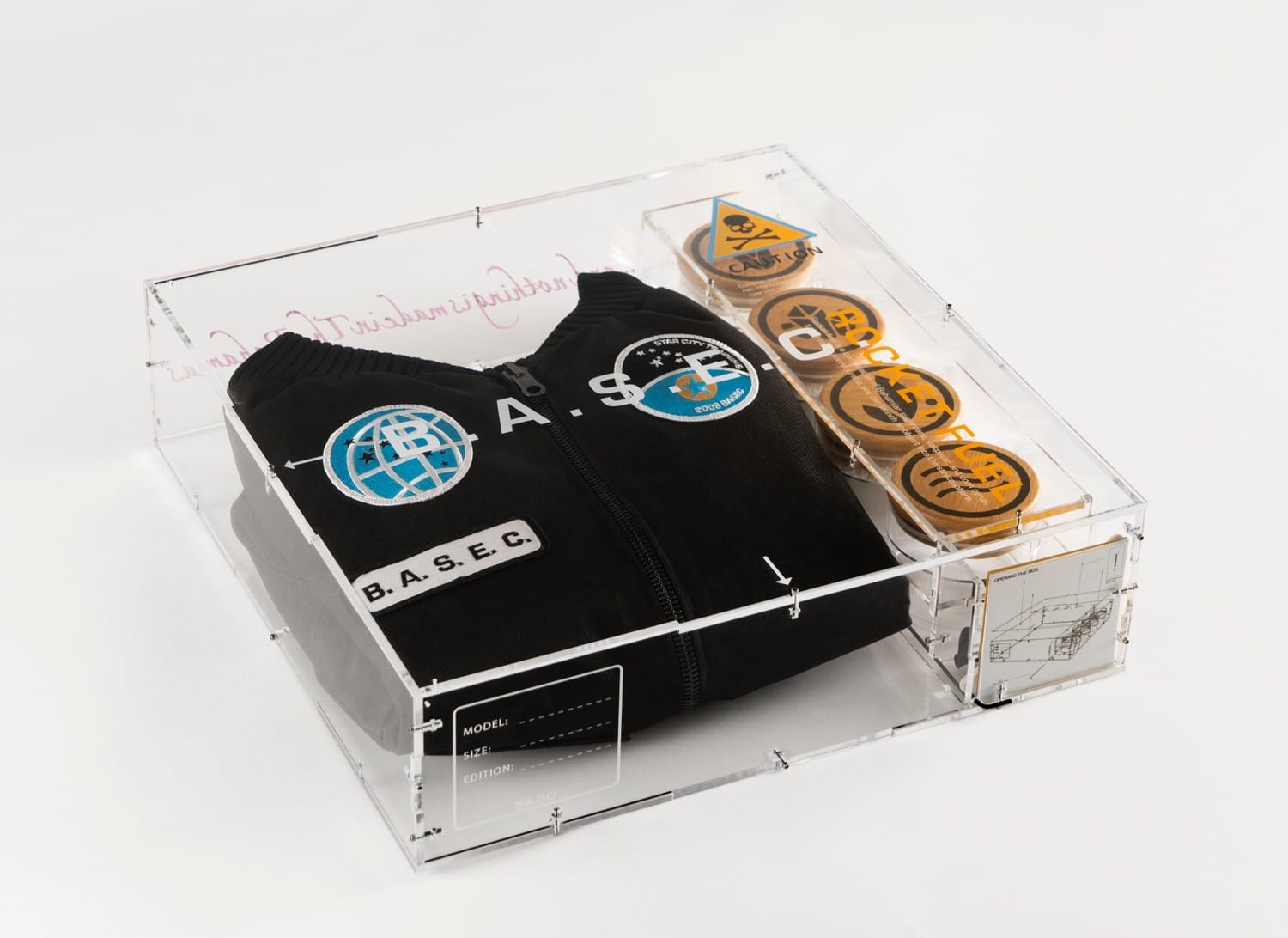 Black hoodie with B.A.S.E.C. logos in a clear display box, next to five circular patch badges with various designs.