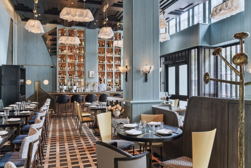 Elegant restaurant interior with modern decor, tall shelves of bottles, cozy seating, and large windows allowing natural light.