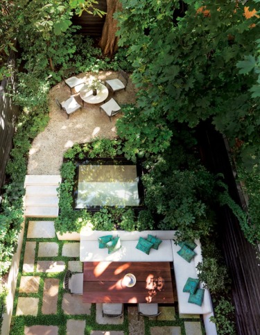 Aerial view of a lush garden with a dining table, chairs, and a circular seating area under leafy trees.