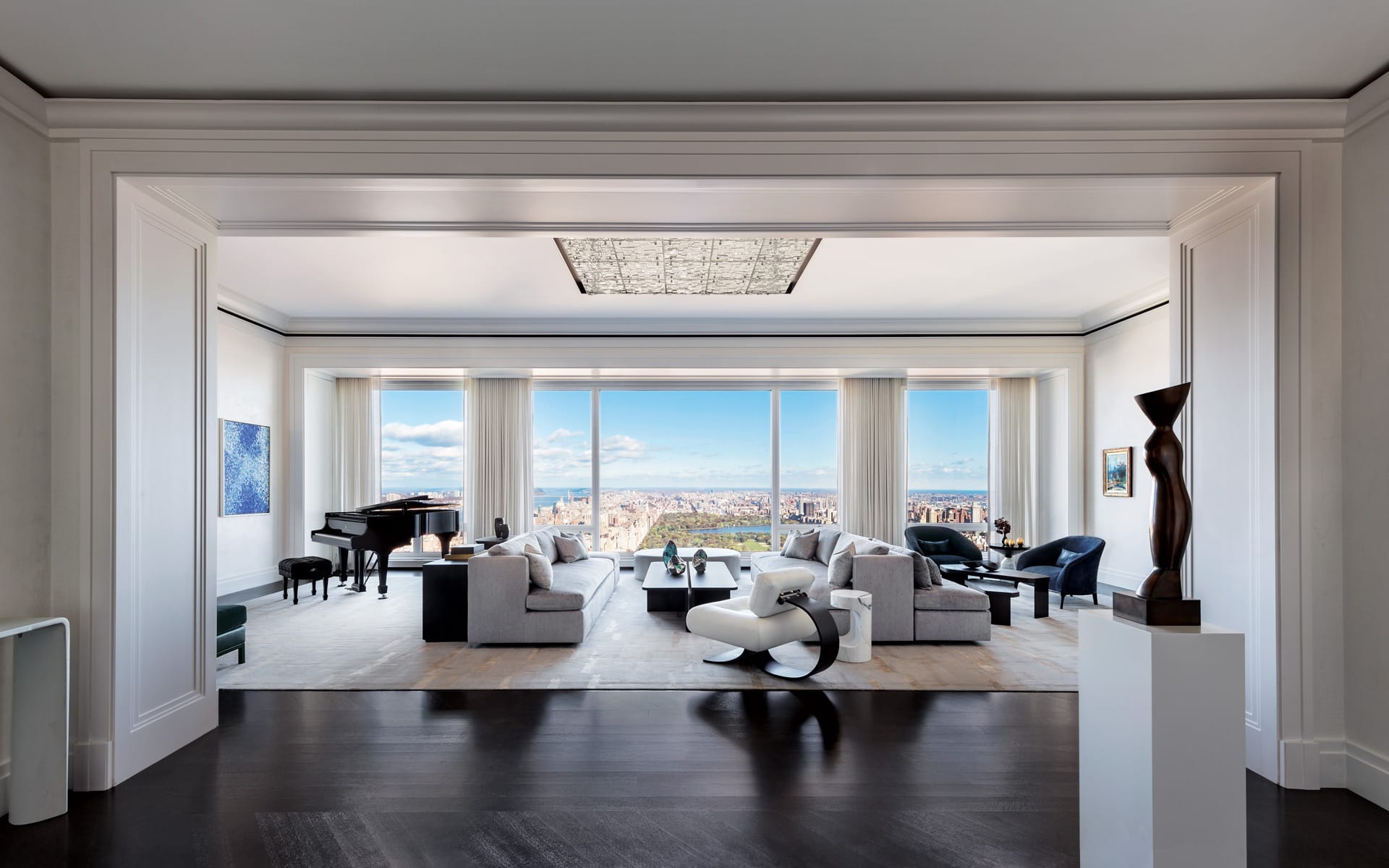 Spacious modern living room with large windows offering a city view, featuring sofas, art pieces, and a grand piano.