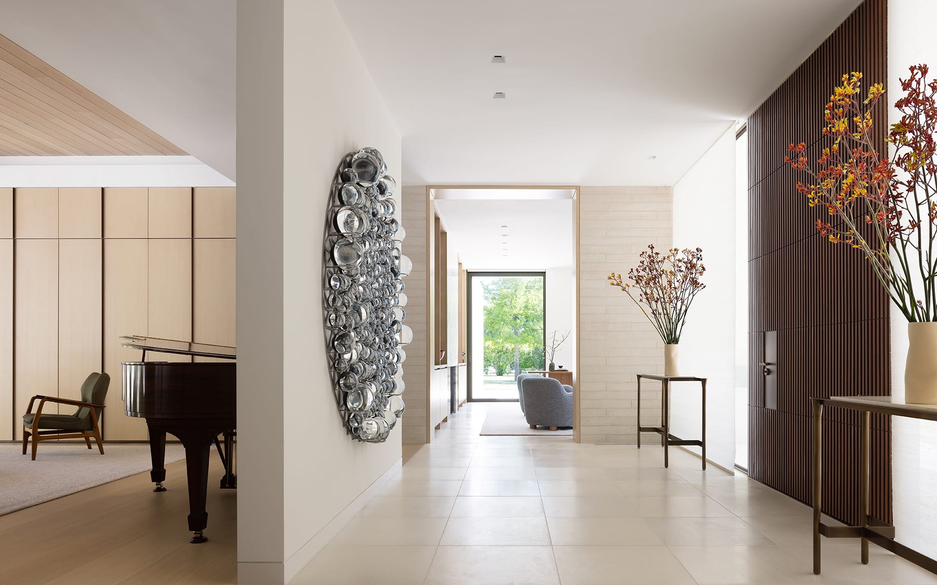 Modern interior hallway with art on wall, wooden accents, grand piano in adjacent room, and vase with flowers by entrance.