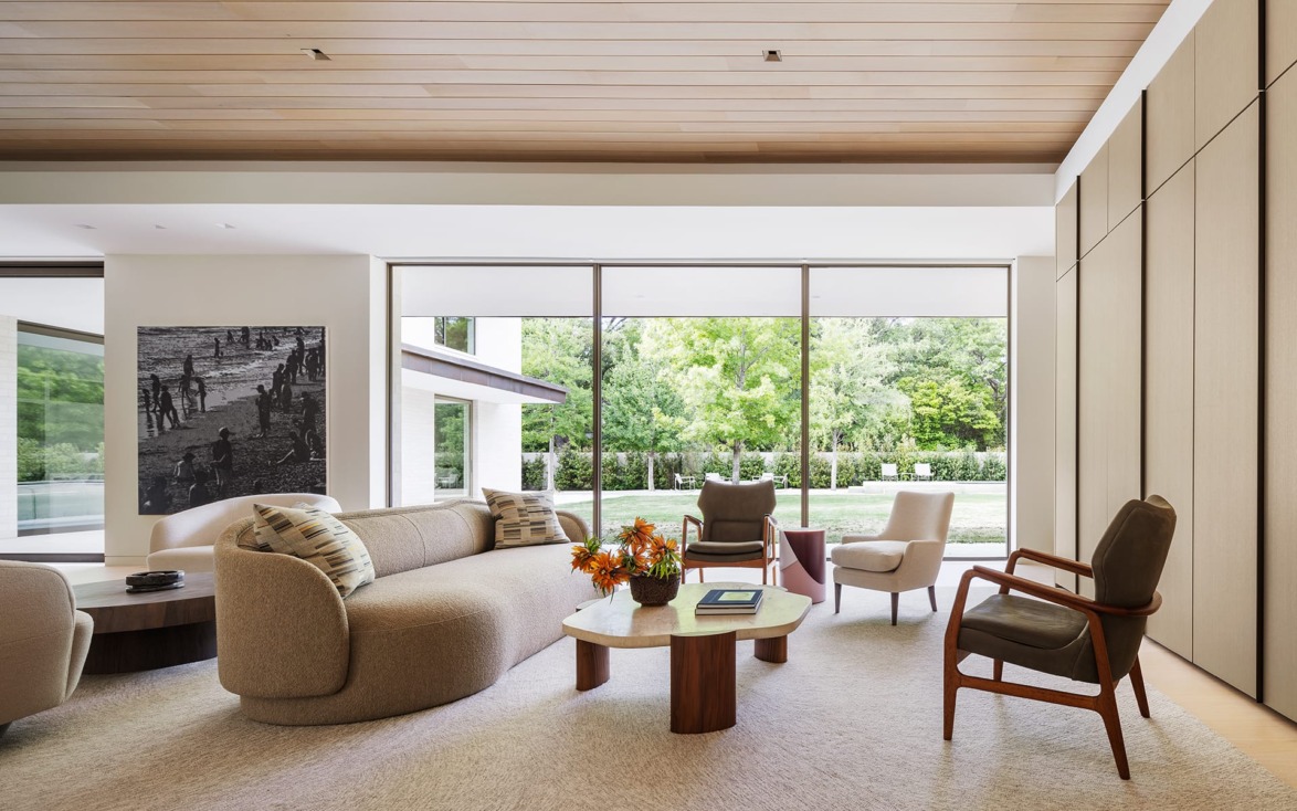 Modern living room with large windows, beige sofa, armchairs, coffee table, and view of garden.