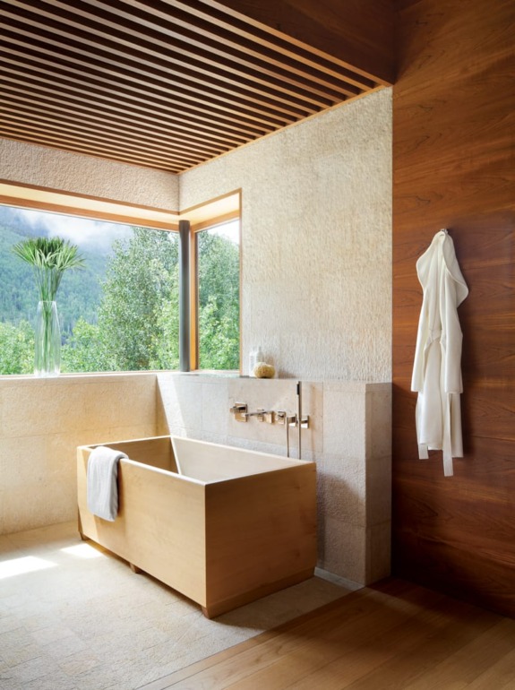 Modern bathroom with a wooden tub, towel on edge, robe hanging, large window view of greenery and mountains.