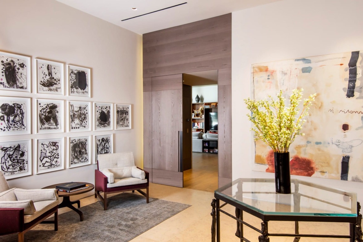 Modern living room with abstract art, two armchairs, a glass table, and a vase with yellow flowers.