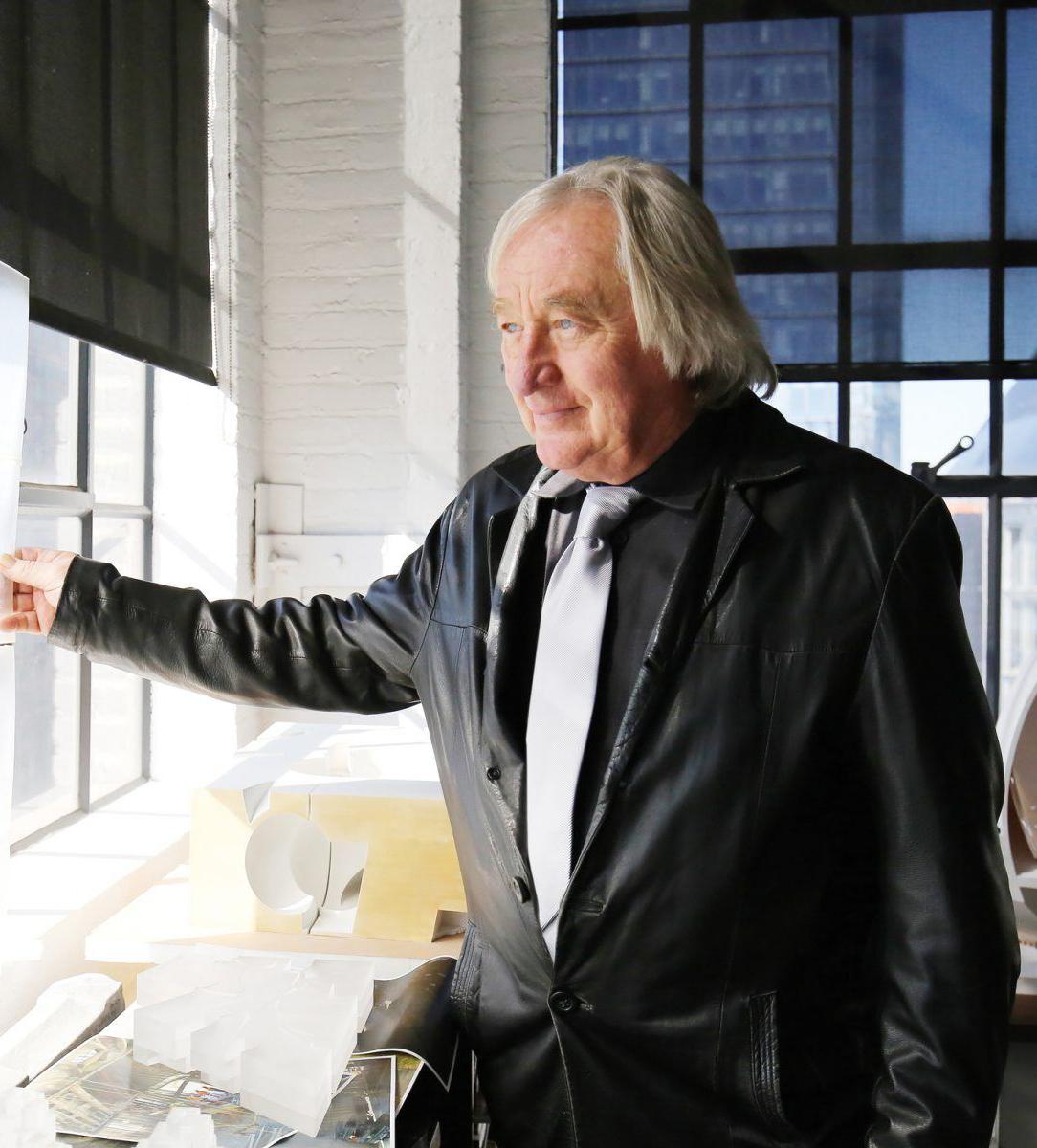 Man in black leather jacket gazing out of a window in a bright office setting with city buildings in the background.
