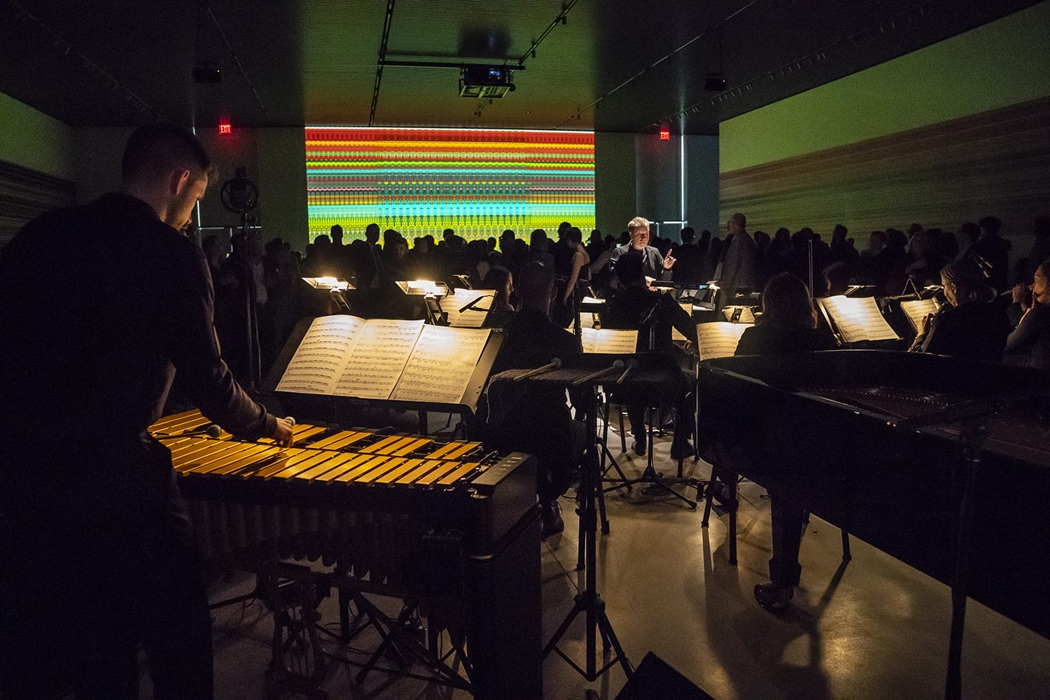 Orchestra performing in dimly lit room with vibrant light display in the background, featuring a percussionist in the foreground.