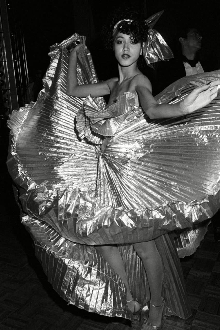 Person wearing an elaborate, shiny costume with wide skirts, striking a dynamic pose in a dimly lit room.