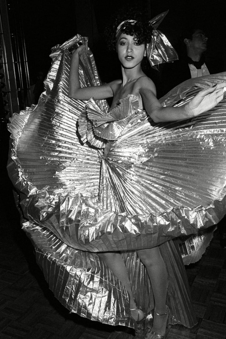 Dancer in a shimmering, pleated metallic costume performing with expressive movement in a dimly lit setting.