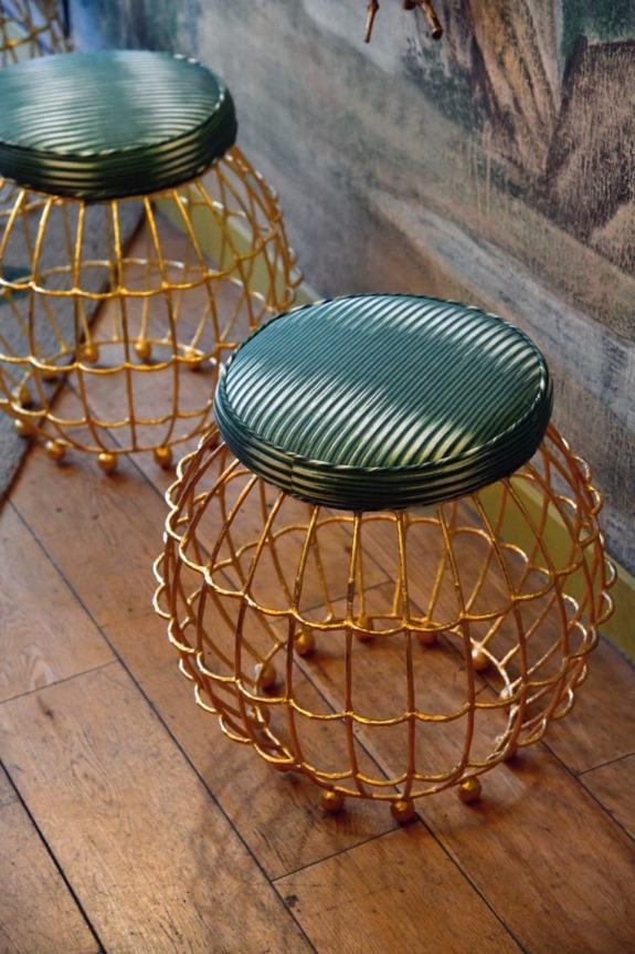 Two stylish stools with green cushioned seats and gold wire frames on a wooden floor in a modern interior setting.
