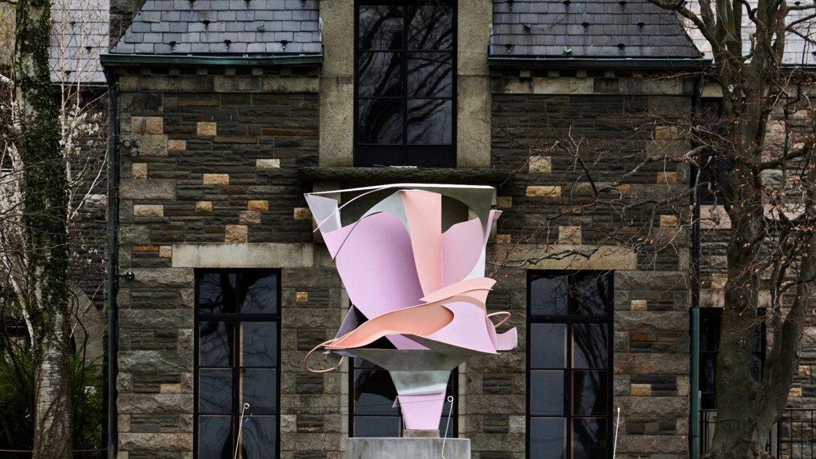 Abstract pink and silver sculpture in front of a stone building with arched windows and a slate roof.