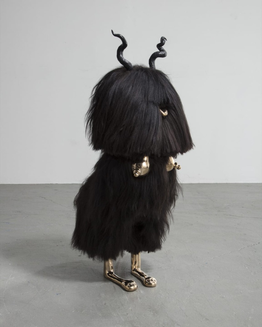 Furry abstract sculpture with black hair and bronze legs, featuring horn-like structures on a plain concrete floor.