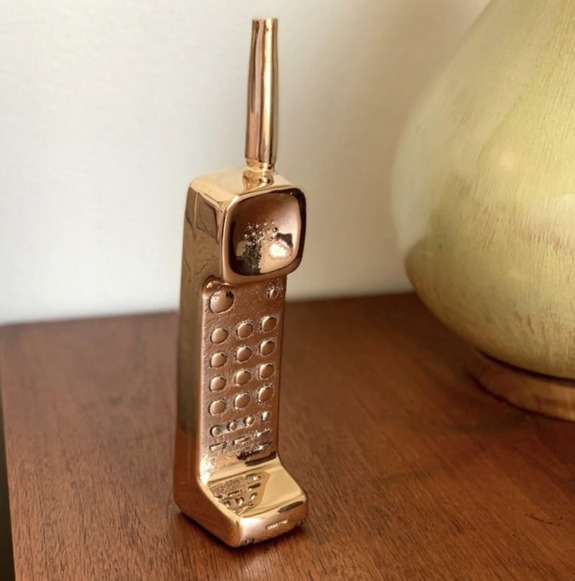 Vintage-style gold-plated mobile phone figurine on a wooden surface next to a light-colored lamp.
