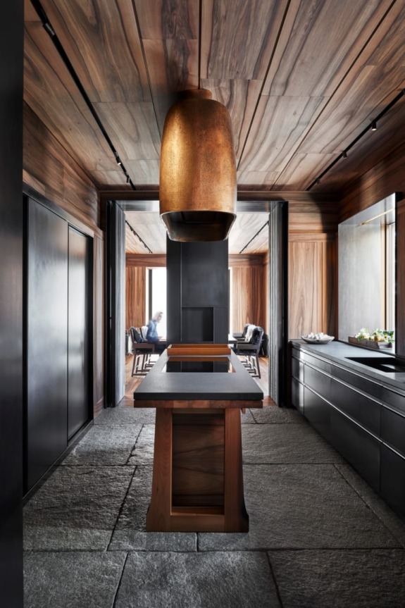 Modern kitchen interior with wooden ceiling, stone flooring, sleek black cabinets, and a large bronze pendant light.