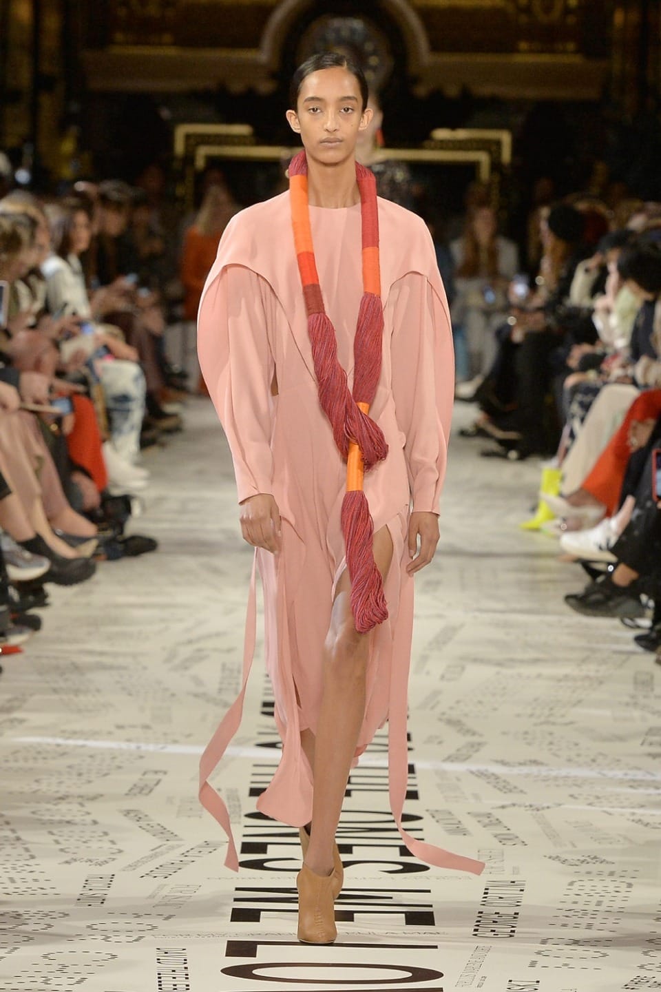 Model walking down a fashion runway in a pink outfit with orange accents, surrounded by an audience.