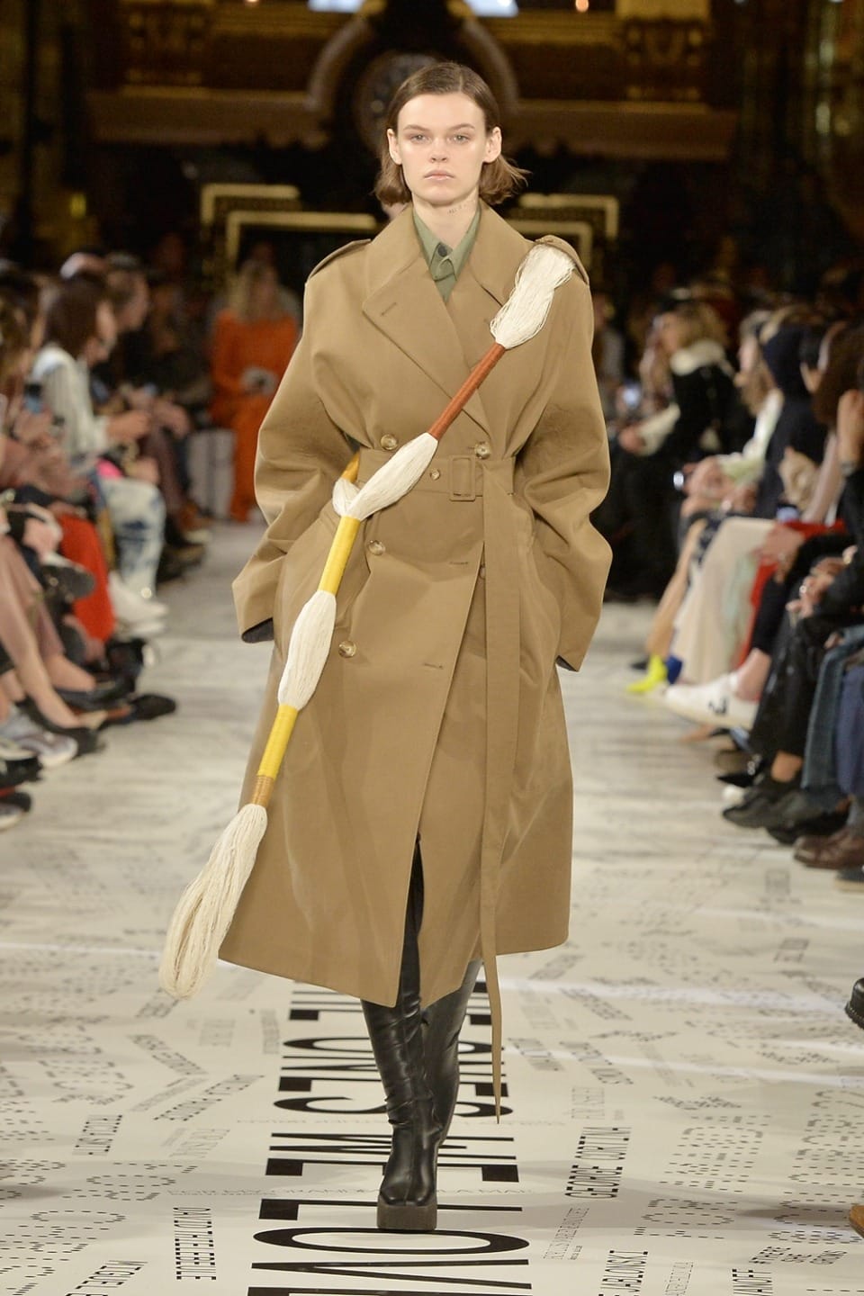 Model walking runway in long tan trench coat and holding a rope-adorned staff, surrounded by seated audience.