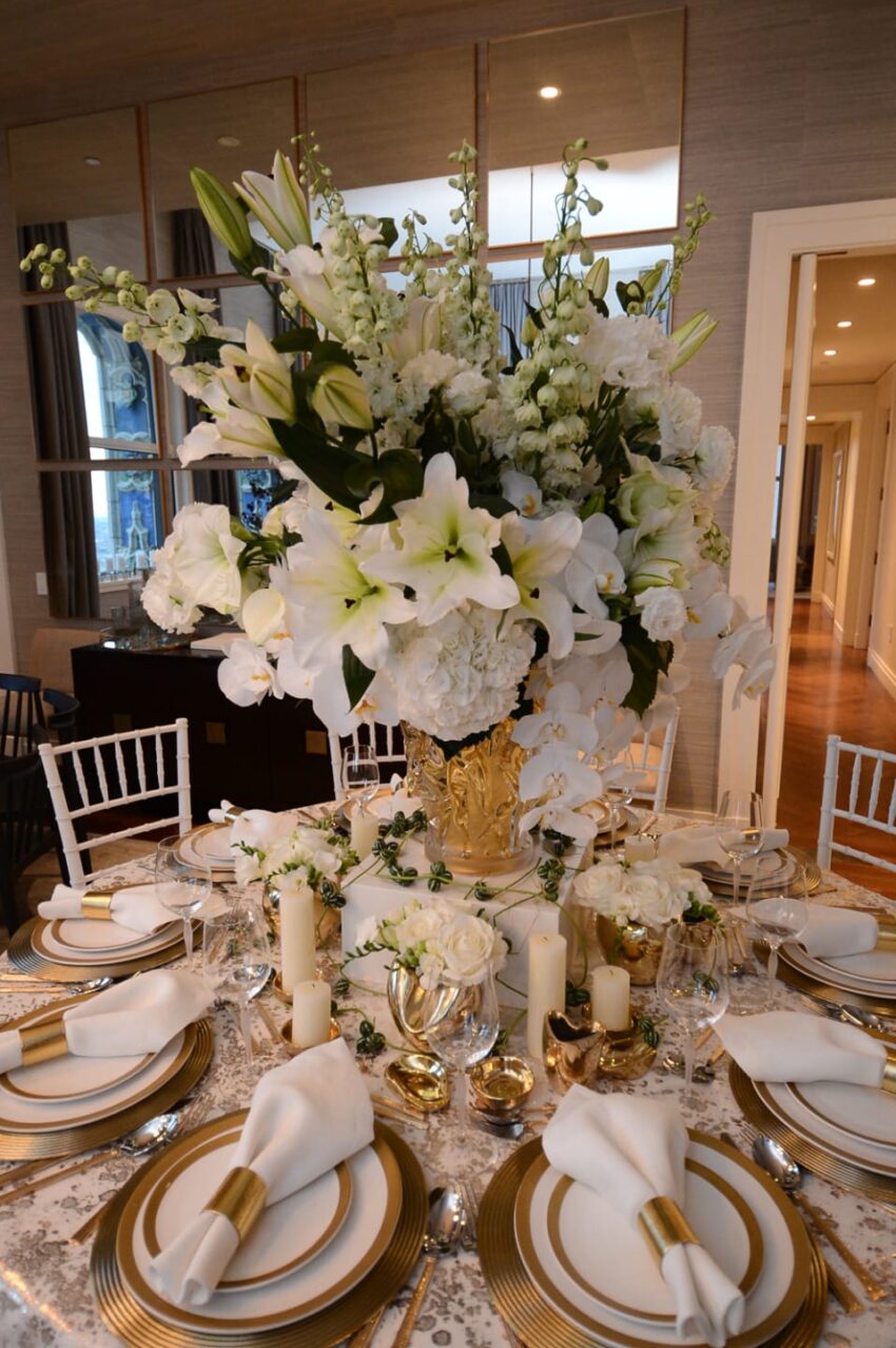 Elegant dining table with large white floral centerpiece, gold accents, white plates, napkins, and candles in a luxurious setting.
