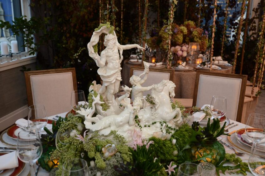 Elegant table setting with ornate white marble centerpiece surrounded by lush greenery and wine glasses in a sophisticated dining room.