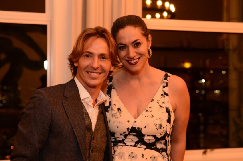 Man in a suit and woman in a floral dress smiling indoors with dim lighting.