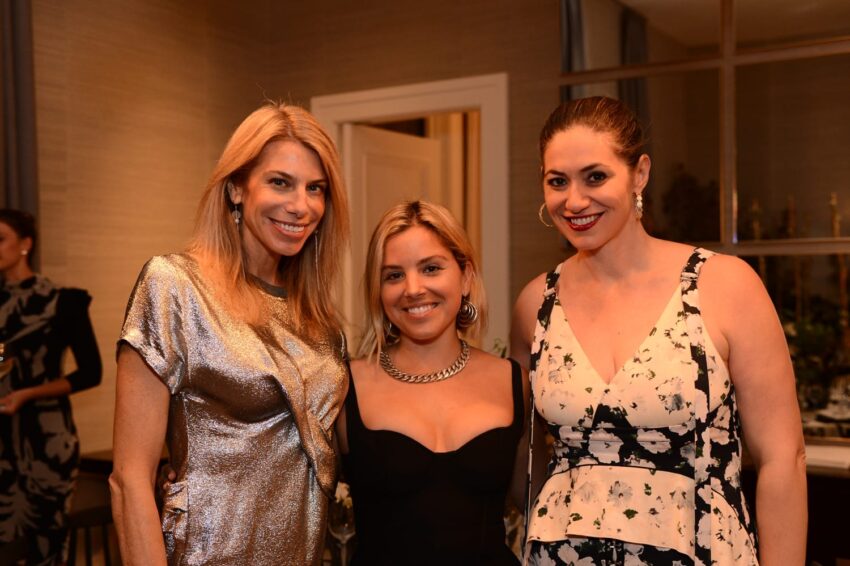 Three women smiling, posing together in a warmly lit room with elegant decor.