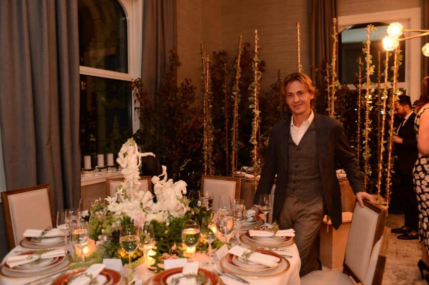 Man standing beside an elegantly set dining table with decorative centerpiece and dimly lit background.