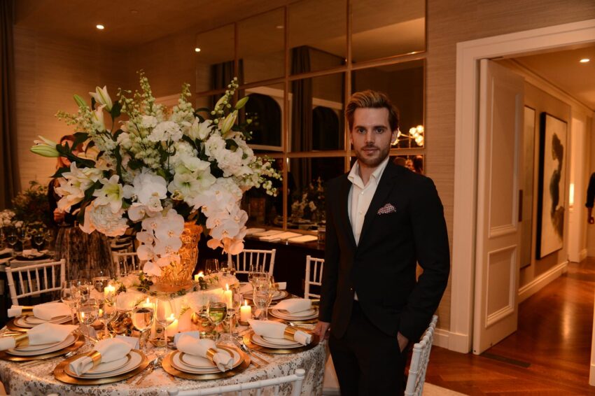 A man in a black suit stands beside an elegantly set dining table with a large floral centerpiece in a luxurious room.