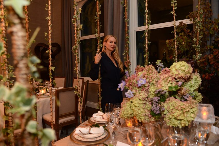 Elegantly dressed woman standing beside a dinner table with floral centerpiece and dimly lit candles in a cozy setting.