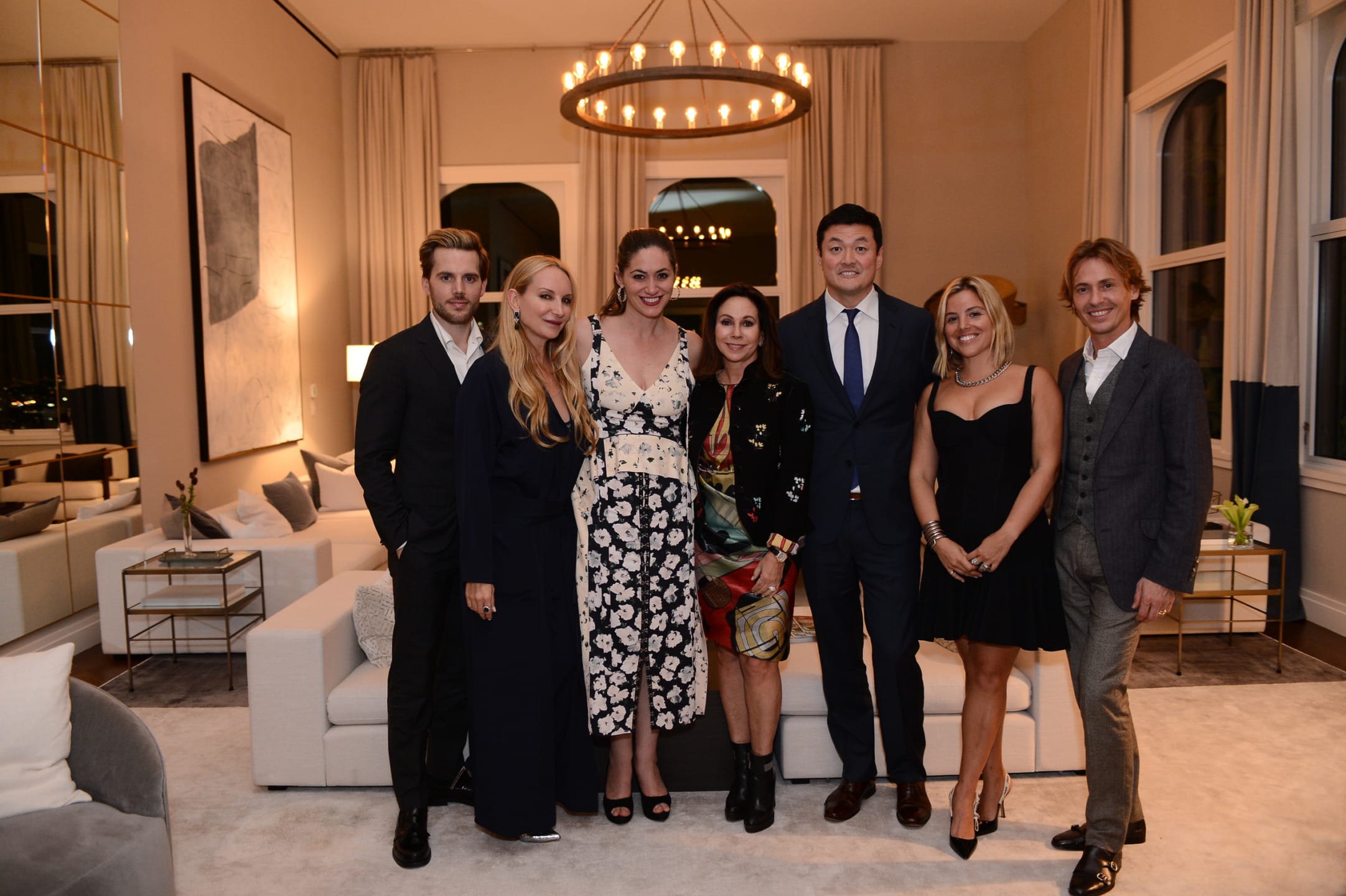 Group of seven people posing in a stylish living room with modern decor and large chandelier.