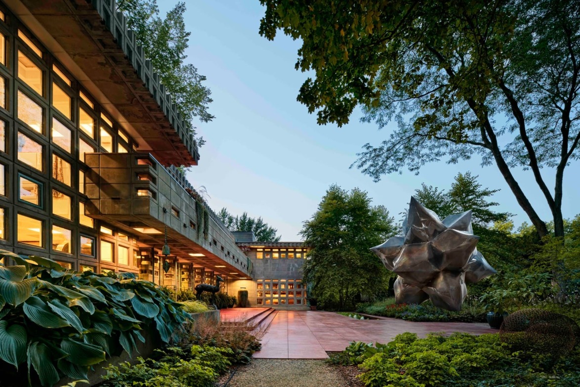 Modern building with large windows surrounded by lush greenery and a metallic sculpture in the courtyard.