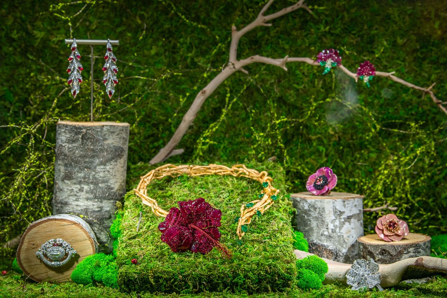 Jewelry display with necklace, earrings, and decorative flowers on moss and wooden logs against a lush green background.