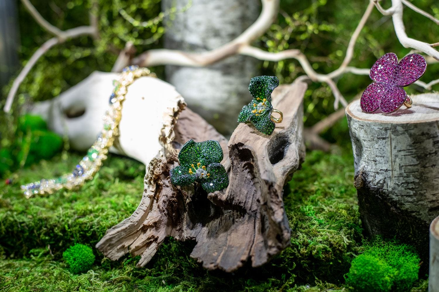 Jewelry with green and pink floral designs displayed on mossy wooden logs in a forest-themed setting.