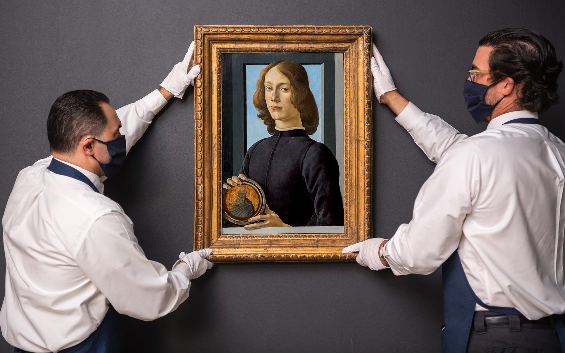 Two gallery workers wearing gloves and masks hang a framed classical portrait on a dark wall