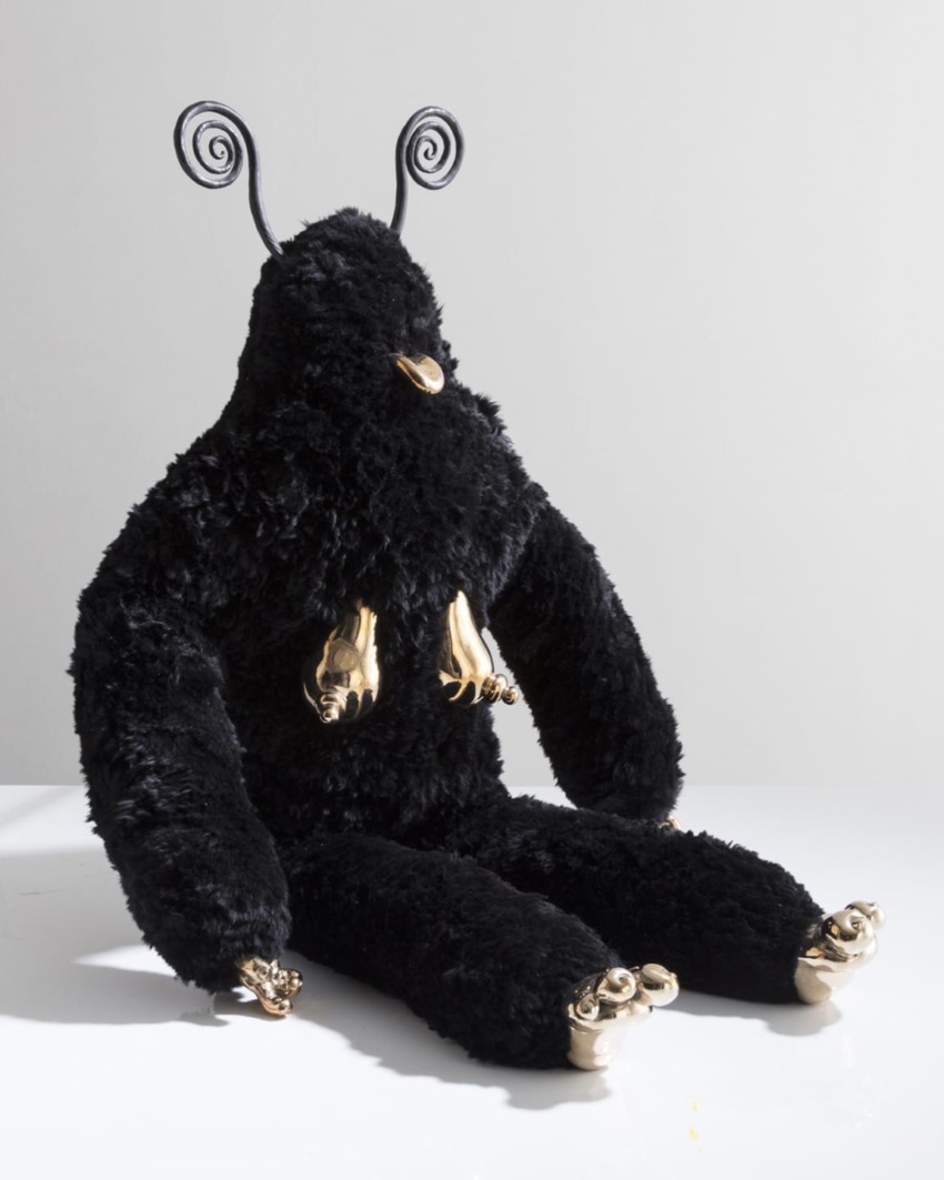 Fluffy black plush creature with spiral antennae, gold nose, and golden hands and feet sitting on a white surface.
