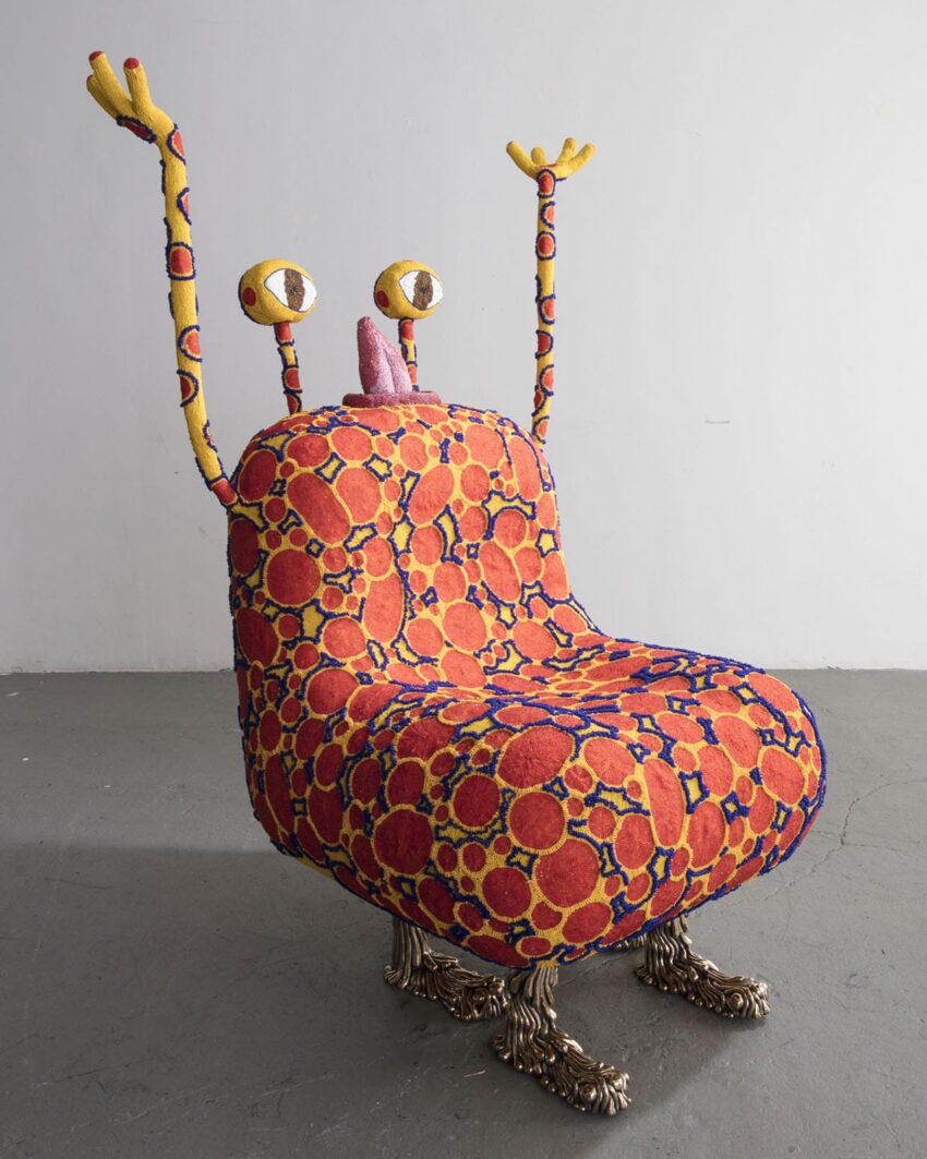 Colorful monster-shaped chair with eye stalks, raised arms, and patterned fabric on bronze-like feet in a minimalist room.