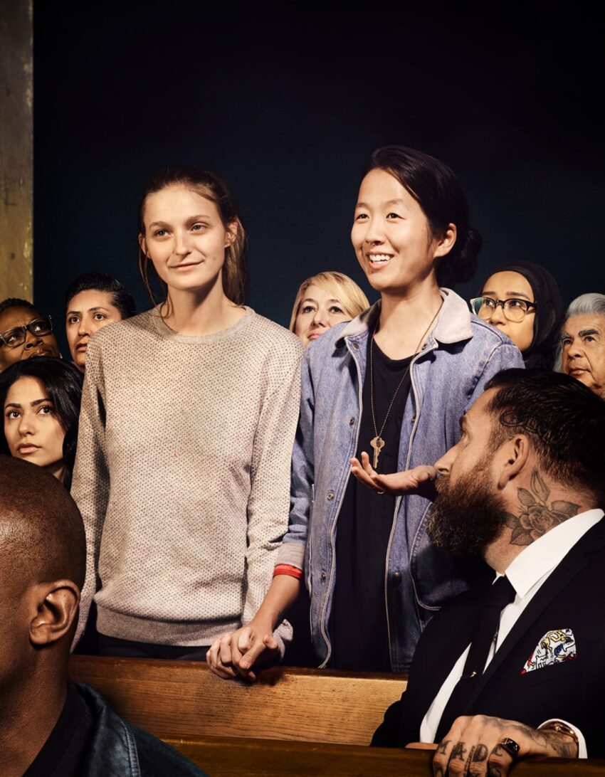 A diverse group of people in a gathering, with two women holding hands, one speaking and others listening attentively.